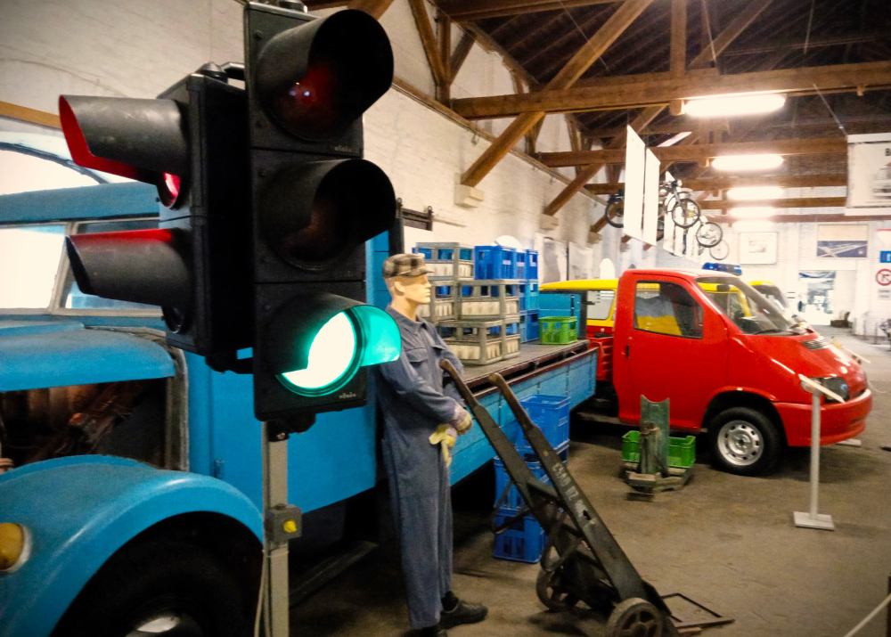 na obrazku sa nachadza semafor s dvomi nakladnymi autami a figurinou robotnika v doprave